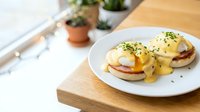 Eggs Benedict with hollandaise sauce served on toasted English muffins with poached eggs, ham, and fresh herbs on white plate for brunch, breakfast, or culinary content.