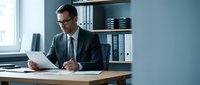 Professional businessman in suit and glasses reviewing documents at modern office desk with organized shelves and natural lighting for corporate presentations.