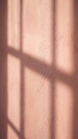 Soft window shadows cast geometric patterns on textured pink wall surface creating warm natural lighting atmosphere for interior design projects.