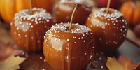 Caramel coated apples decorated with white sprinkles on dark background with autumn leaves and pumpkins, traditional fall season sweet treat.