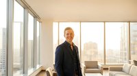 Professional middle-aged Caucasian businessman in dark suit standing confidently in modern office with floor-to-ceiling windows overlooking city skyline.