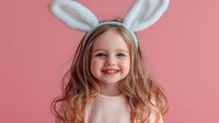 Happy little girl wearing white bunny ears smiling at camera against pink background for Easter celebration and spring holiday festivities.