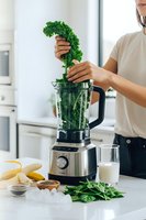 Young woman adding fresh kale to blender for healthy green smoothie preparation in modern kitchen with organic ingredients and nutritious lifestyle concept.