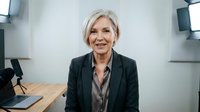 Mature woman with white hair and professional attire smiling at camera in modern office studio setting with laptop and lighting equipment, suitable for business and corporate content.