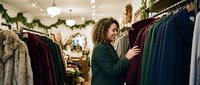 Woman with curly brown hair and medium skin tone browsing clothing racks in a boutique shop decorated with holiday garlands and warm lighting.