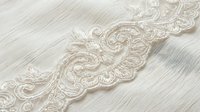 Ornate ivory lace trim with intricate scrollwork and floral embroidery on white fabric background, suitable for wedding dress design, bridal accessories, and textile decoration projects.