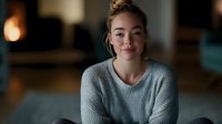 Young Caucasian woman with blonde hair in casual gray sweater smiling warmly at camera in cozy modern living room with soft lighting and blurred background.