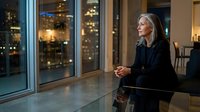 Mature woman with long gray hair, wearing black blazer, standing by floor-to-ceiling windows overlooking illuminated city skyline at night. Professional business setting, corporate office environment, evening ambiance with warm interior lighting.