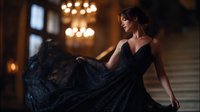 Young Caucasian woman in flowing black evening gown dancing gracefully in luxurious interior with chandeliers and grand staircase. Romantic fashion portrait in moody lighting.