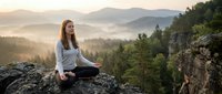 Young woman meditating on mountain rock at sunrise with misty valley landscape. Peaceful mindfulness practice in nature for wellness and mental health.