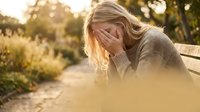 Young blonde woman sitting on park bench covering face with hands in emotional distress during golden hour sunset light for mental health awareness.