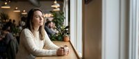 Young woman sitting by cafe window with coffee cup, contemplating while looking outside. Cozy restaurant interior with warm lighting and blurred customers.