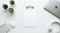 Clean white workspace with blank clipboard, laptop, keyboard, mouse, headphones and small potted plants on desk for business planning and productivity.