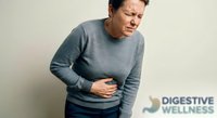 Middle-aged Asian man experiencing severe stomach pain holding abdomen in discomfort wearing casual gray sweater against neutral background for health concepts.