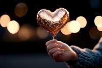 Heart-shaped lollipop held by hand against blurred bokeh lights background creates romantic mood, warm evening atmosphere with golden glow and sparkles.