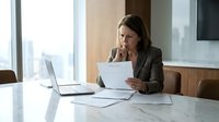 Mature woman with brown hair, Caucasian ethnicity, wearing business attire, reviewing documents at marble desk with laptop in modern office with city skyline view.