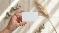 Female hand holding blank white business card mockup with natural shadows from dried plants on neutral beige background for branding design.