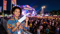 Young woman with curly hair and medium skin tone holding ticket at night music festival with illuminated stage, crowd, and colorful lights for entertainment and event promotion.