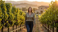 Young woman walking through vineyard rows at golden hour sunset with mountains and winery in background for wine tourism and agricultural lifestyle content.