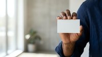 Man in dark blue shirt holding blank white business card, isolated on blurred office background for corporate branding and professional networking purposes.