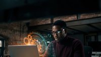 African American male developer working on laptop with holographic data visualization and digital interface elements in modern tech workspace.