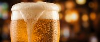 Cold beer glass with golden lager and thick foam head, condensation droplets on glass, blurred warm bokeh background, suitable for beverage, pub, and hospitality marketing.