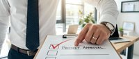 Professional businessman in white shirt and tie marking project approval checkbox on document at modern office desk with laptop and plants in background.