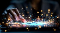 Finger touching digital screen with magical glowing particles and light effects creating futuristic interactive experience on dark background.
