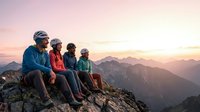 Group of four diverse hikers sitting on mountain summit at sunset, enjoying panoramic alpine landscape view. Outdoor adventure and friendship concept for travel and lifestyle use.