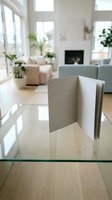 Blank open greeting card standing on glass table in bright modern living room with fireplace and plants, mockup for design.