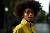 Young African American woman with natural curly afro hair wearing bright yellow sports jacket looking confidently at camera against blurred urban background.