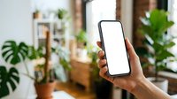 Young adult with dark skin tone holding smartphone with blank white screen in modern home interior with potted plants and natural lighting for app mockup or mobile technology promotion.