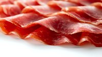 Thinly sliced prosciutto ham arranged in wavy rows showing delicate marbling and texture on white background, macro food photography for culinary and gastronomy use.