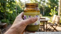 Hand holding mason jar with green smoothie drink outdoors in sunny garden setting with natural bokeh background for healthy lifestyle content.