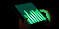 Person using tablet in dark environment with glowing green bar graph display, finger touching screen to analyze financial data or statistics.