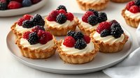 Fresh berry tartlets with smooth vanilla custard filling topped with ripe raspberries and blackberries on flaky golden pastry shells, served on white plate.