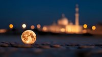Full moon rising near mosque at dusk, glowing orange orb resting on sandy beach with blurred religious architecture and city lights in background.