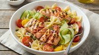 Grilled chicken breast slices over fresh pasta salad with cherry tomatoes, cucumber ribbons and herbs on rustic wooden table for healthy dining.