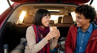 Young multiethnic couple enjoying takeout food in car trunk during road trip adventure, sharing meal and laughing together in cozy vehicle setting.