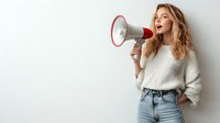 Young blonde woman shouting into red and white megaphone against white background. Casual outfit with sweater and jeans for communication concepts.