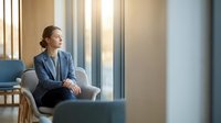 Professional businesswoman in blue suit sitting thoughtfully in modern office chair by large windows with natural lighting for corporate presentations.