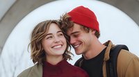 Young romantic couple laughing together outdoors, man wearing red beanie and woman in burgundy sweater sharing intimate moment against white background.