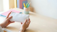 Female hands holding blank white envelope at wooden desk with colorful stationery, office supplies and books for business communication and correspondence projects.