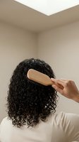 Woman with curly dark hair using wooden brush for daily hair care routine in modern bathroom setting for beauty and wellness concepts.