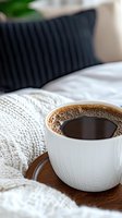 Fresh black coffee in white ceramic cup on wooden tray with cozy knit blanket and dark pillow background, creating relaxing morning atmosphere.