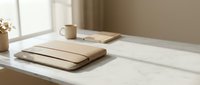 Minimalist workspace with stacked notebooks and coffee cup on white marble desk by window with natural sunlight creating warm shadows and peaceful morning atmosphere.