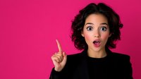 Young mixed race woman with curly hair showing surprised expression and pointing finger up against bright pink background. Black blazer, natural makeup.