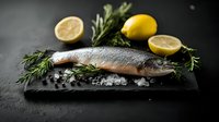 Raw fresh salmon fillet on black stone board with ice, lemon slices, rosemary sprigs and peppercorns against dark background. Food photography.