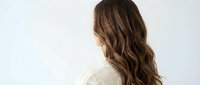 Young woman with long wavy brown hair viewed from behind wearing white blouse against light background for beauty and lifestyle concepts.