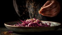 Sliced red onion rings on rustic plate with hand sprinkling spices, dramatic dark food photography against black background.